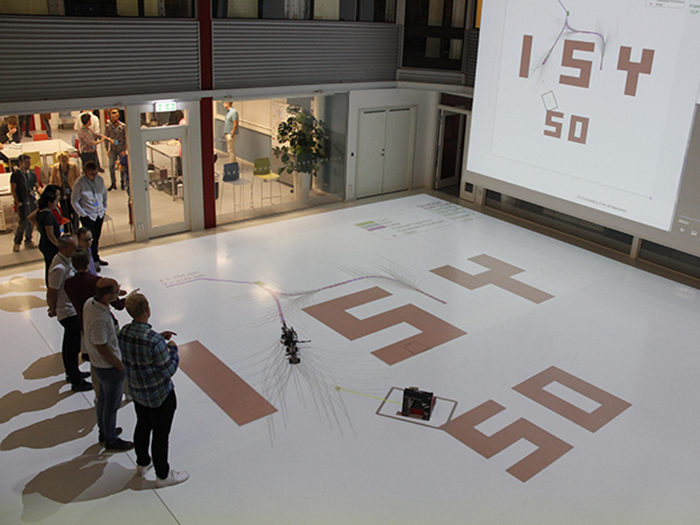 People standing on the floor of the research arena. One person holds a remote control. Projectors are displaying the words "ISY 50" on the flor and one wall. A small vehicle is moving following lines on the floor.